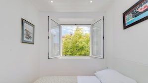 Un dormitorio con una cama, una ventana con contraventanas abiertas y un cuadro en la pared.