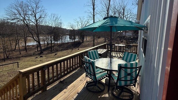 Une terrasse en bois avec une table et des chaises, placée sous un parasol vert, d’où l’on peut admirer un plan d’eau et des arbres.