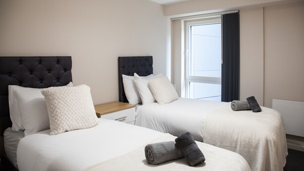 A hotel room with two beds, each with a headboard, white bedding, and a rolled towel on the bed.