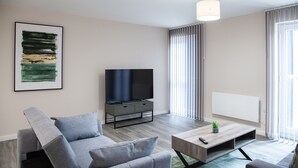 A modern living room with a grey sofa, a flat-screen TV on a wooden stand, and a coffee table with a remote control.
