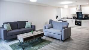 A modern living room with a grey sofa, a grey armchair, and a wooden coffee table. There is a kitchen area with white cabinets and a black microwave.
