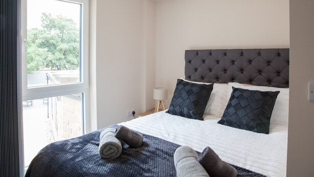 A bedroom with a large window, a tufted headboard, and a bed with rolled towels.