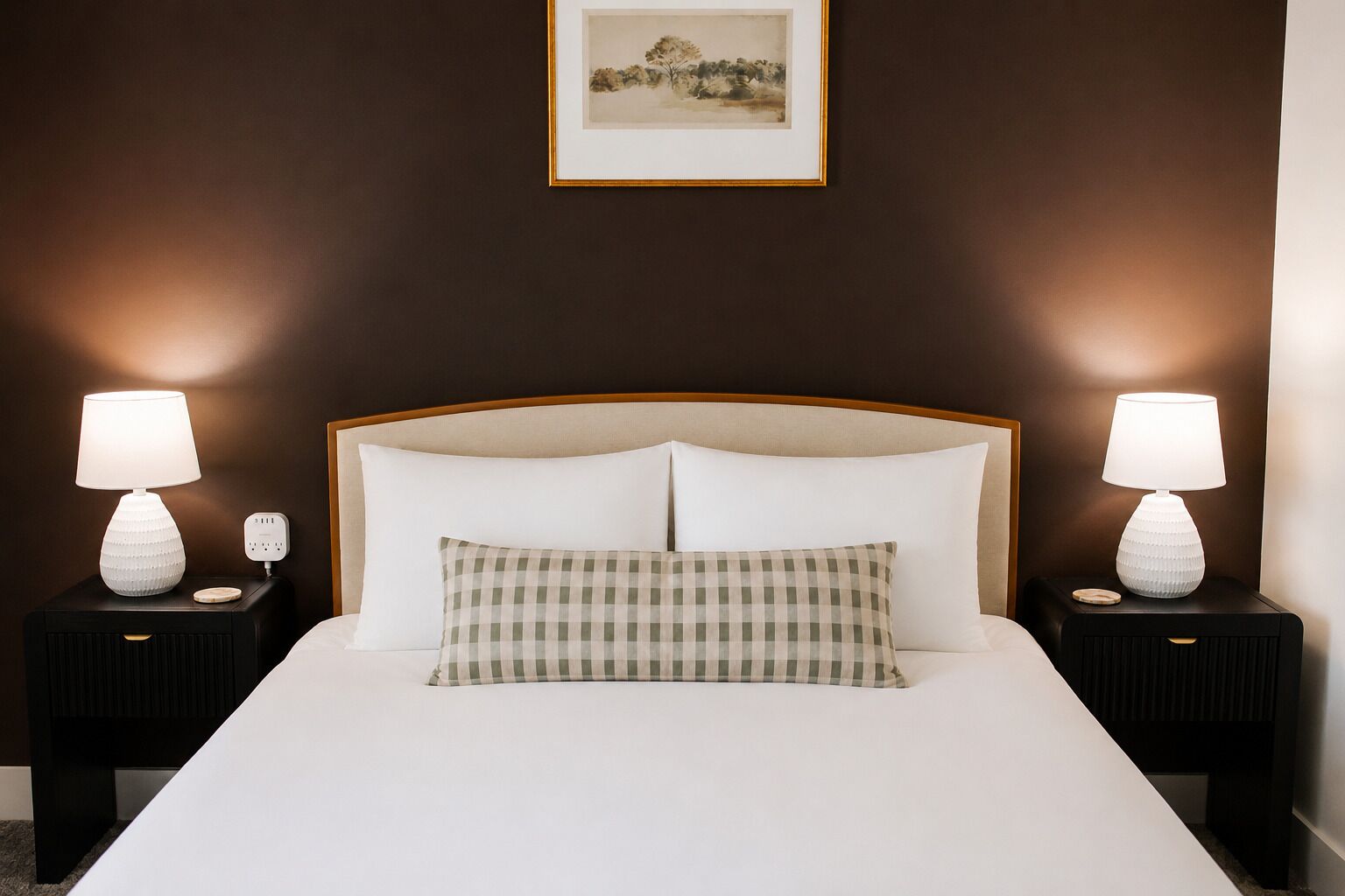 Habitación de hotel con una cama grande, dos mesitas de noche con lámparas, un cojín a cuadros y un cuadro en la pared.