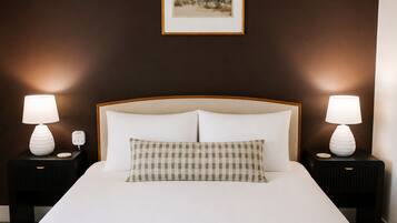 Habitación de hotel con una cama grande, dos mesitas de noche con lámparas, un cojín a cuadros y un cuadro en la pared.