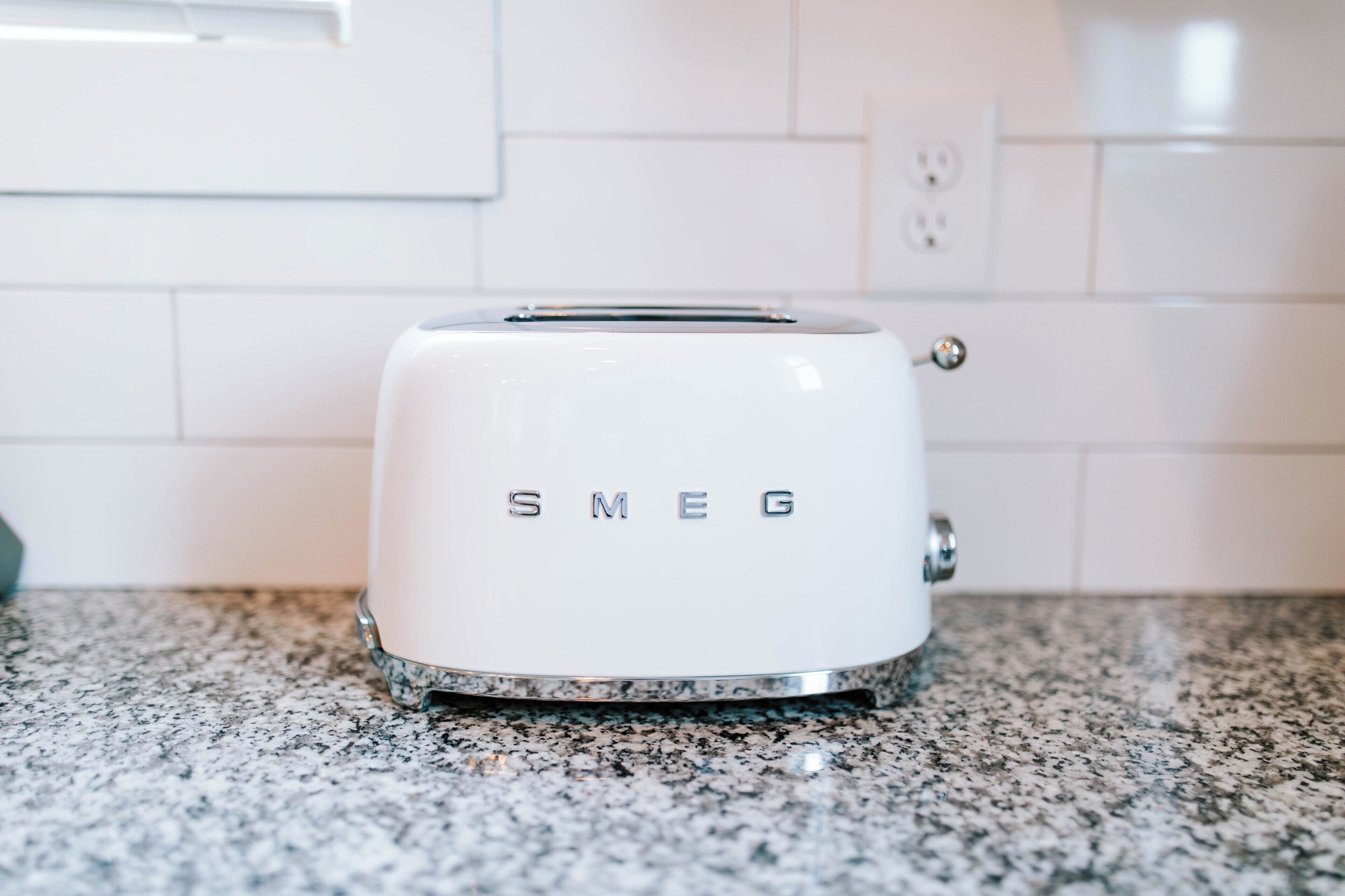 A white Smeg toaster on a granite countertop in a kitchen with white tiled walls.