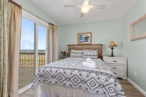 Un dormitorio con cama, mesita de noche, lámpara, ventilador de techo, cuadro enmarcado y vista a la playa por la ventana.