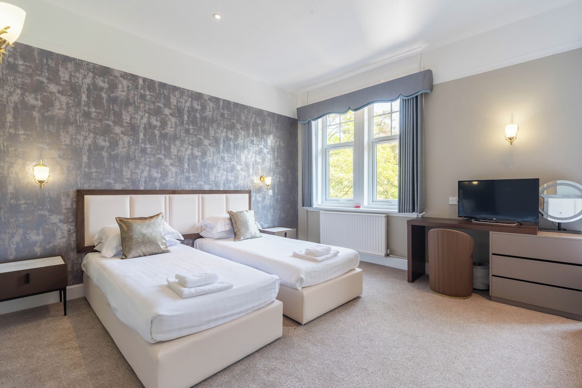 A modern hotel room with two beds, a desk, a TV, and a window with curtains.