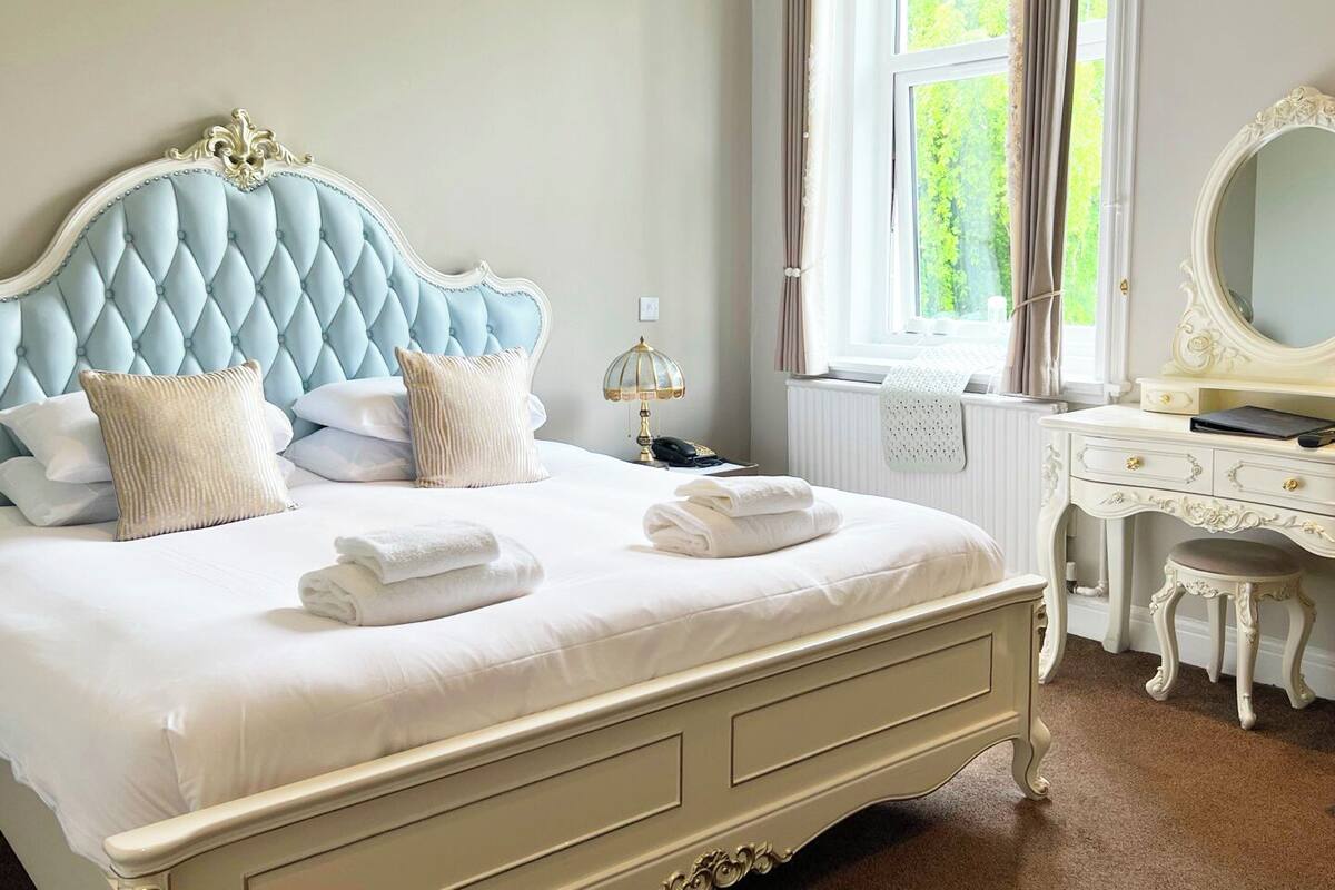 A bedroom with a tufted headboard, a bedside table with a lamp, a dresser, and a window with curtains.