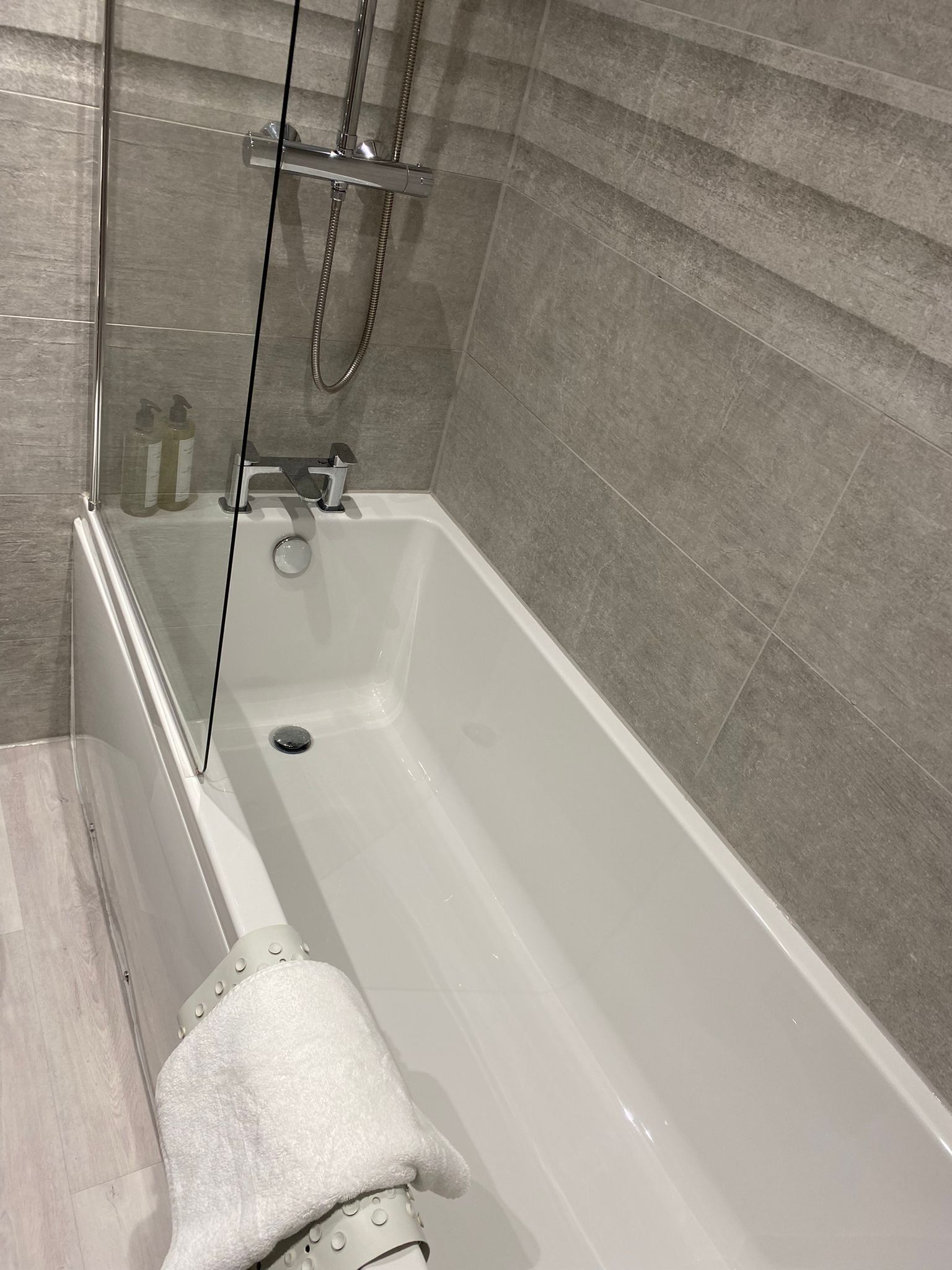 A modern bathroom with a glass-enclosed shower, a white bathtub, and grey tiled walls.