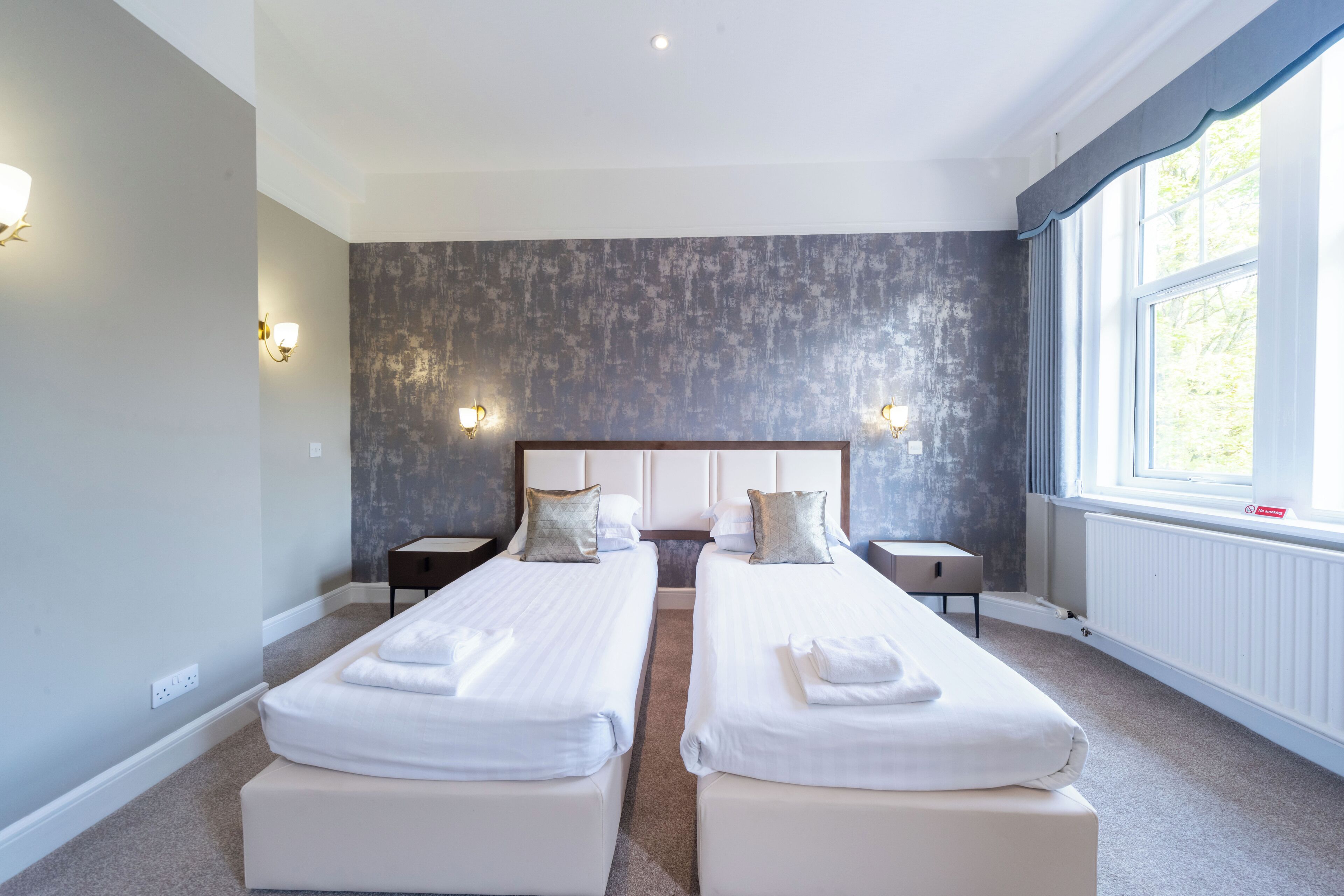 Una habitación de hotel con dos camas individuales, un ventanal amplio y una pared gris con textura.