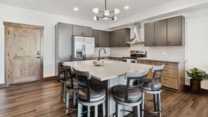A modern kitchen with a central island, bar stools, stainless steel appliances, and wooden flooring.