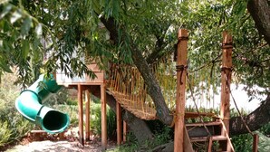 A wooden play structure with a slide and climbing net, surrounded by trees and grass. - Cabañas Elsa María (Atlautla)