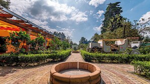 A paved pathway leading to a circular stone structure, flanked by neatly trimmed hedges and a building with an orange facade. - Cabañas Elsa María (Atlautla)