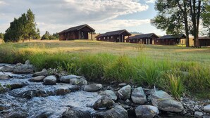 Un chalet rustique doté de plusieurs cabanes, avec un ruisseau au premier plan et une colline verdoyante en arrière-plan.