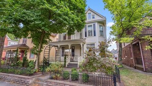 A two-story house with a porch, a small front yard, and a brick building to the right. - Amazing Location! Short North Victorian Oasis 5-bedroom w Hot Tub and Peleton (Columbus)