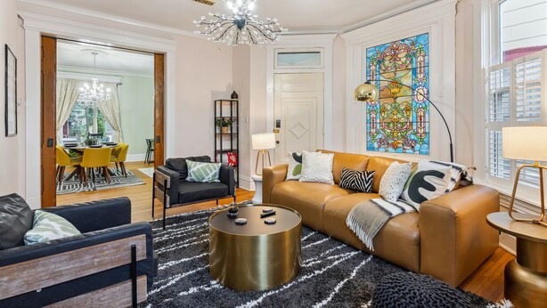 A modern living room with a sofa, coffee table, and chandelier. There is a dining area with yellow chairs and a table visible through an open door. - Amazing Location! Short North Victorian Oasis 5-bedroom w Hot Tub and Peleton (Columbus)