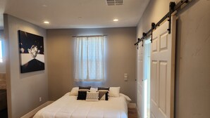 A bedroom with a large bed, a window with sheer curtains, a sliding door, and a wall-mounted picture.