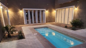 A swimming pool with a tiled deck, surrounded by a textured wall and a window with curtains.