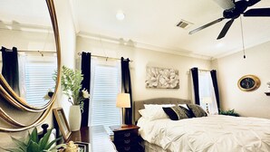 A bedroom with a bed, a ceiling fan, a nightstand, a lamp, a mirror, and a wall clock. - The Sweet Magnolia 4BR Home by VelvetDitchVillas (Oxford)
