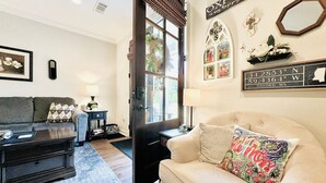 A cozy living room with a sofa, a coffee table, and a lamp. There is a door leading outside, and the room has a window. - The Sweet Magnolia 4BR Home by VelvetDitchVillas (Oxford)