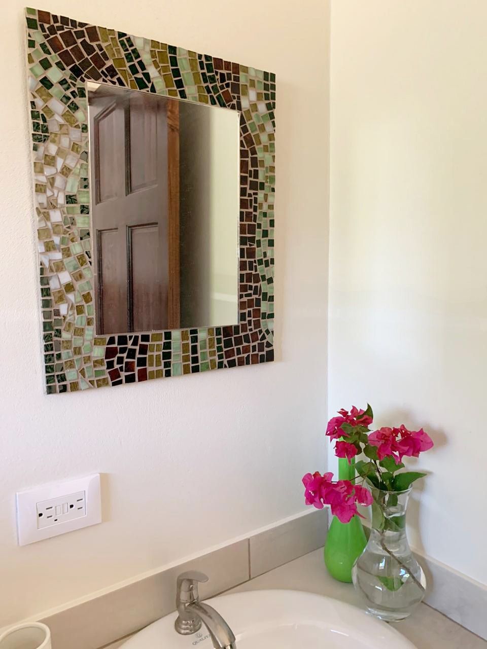 A bathroom with a mosaic tile wall, a mirror, a sink, and a vase with flowers.