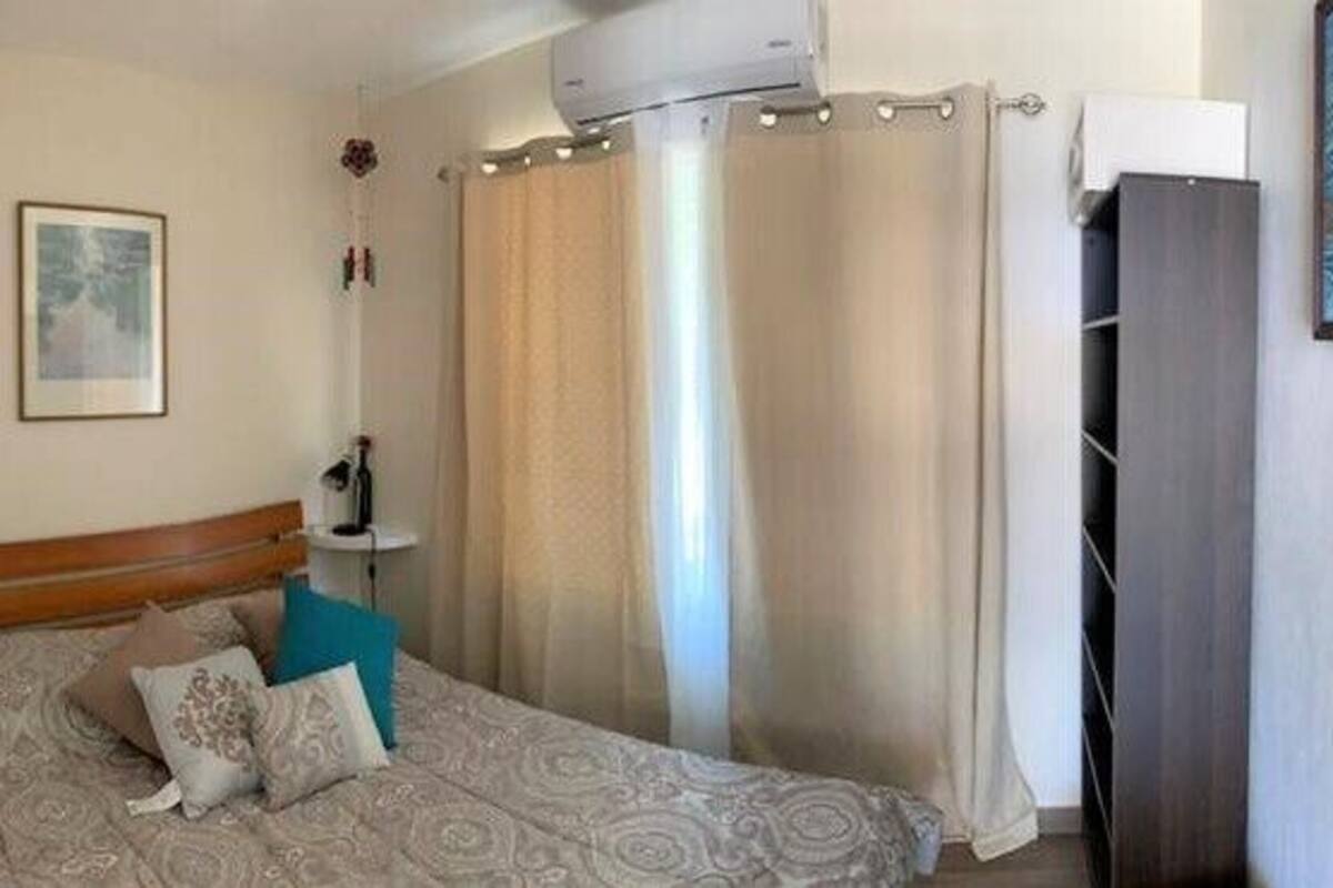 Un dormitorio con cama, cortinas, una mesita de noche e imagen colgada en la pared.