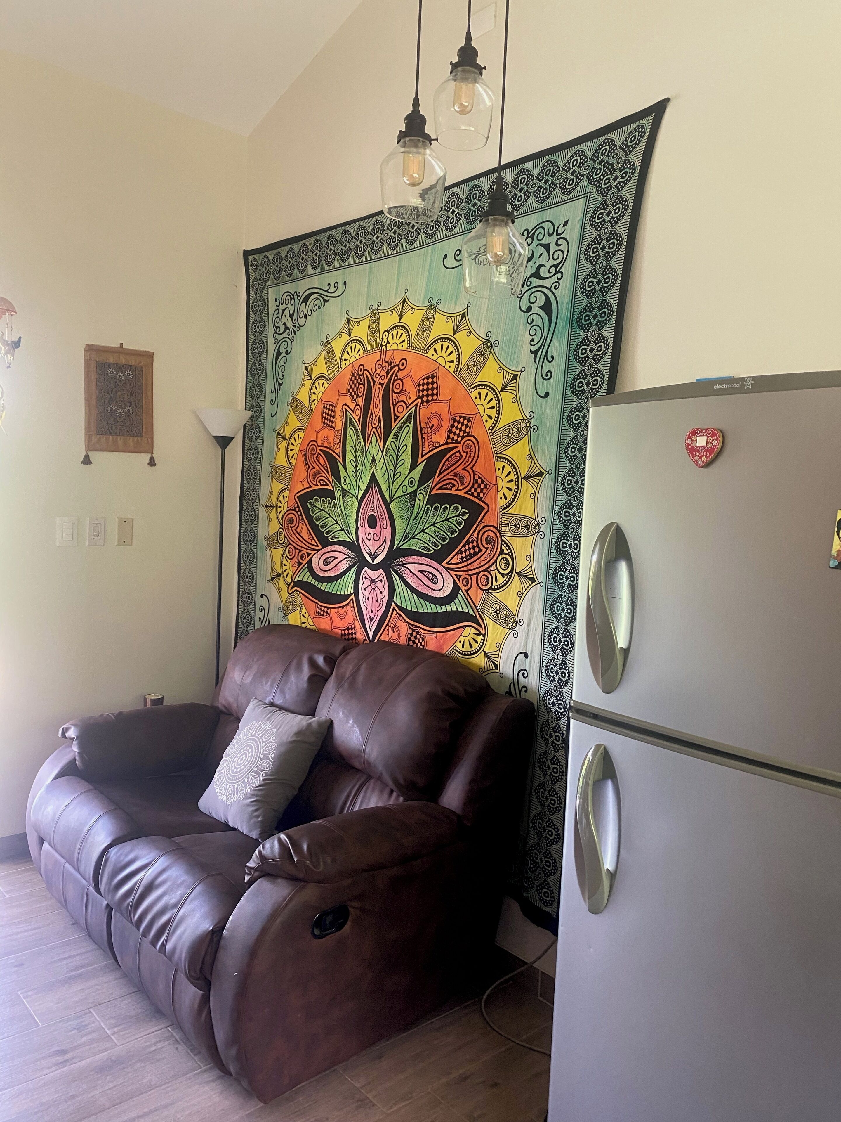 A living room with a brown leather sofa, a patterned tapestry, a modern refrigerator, and pendant lights.