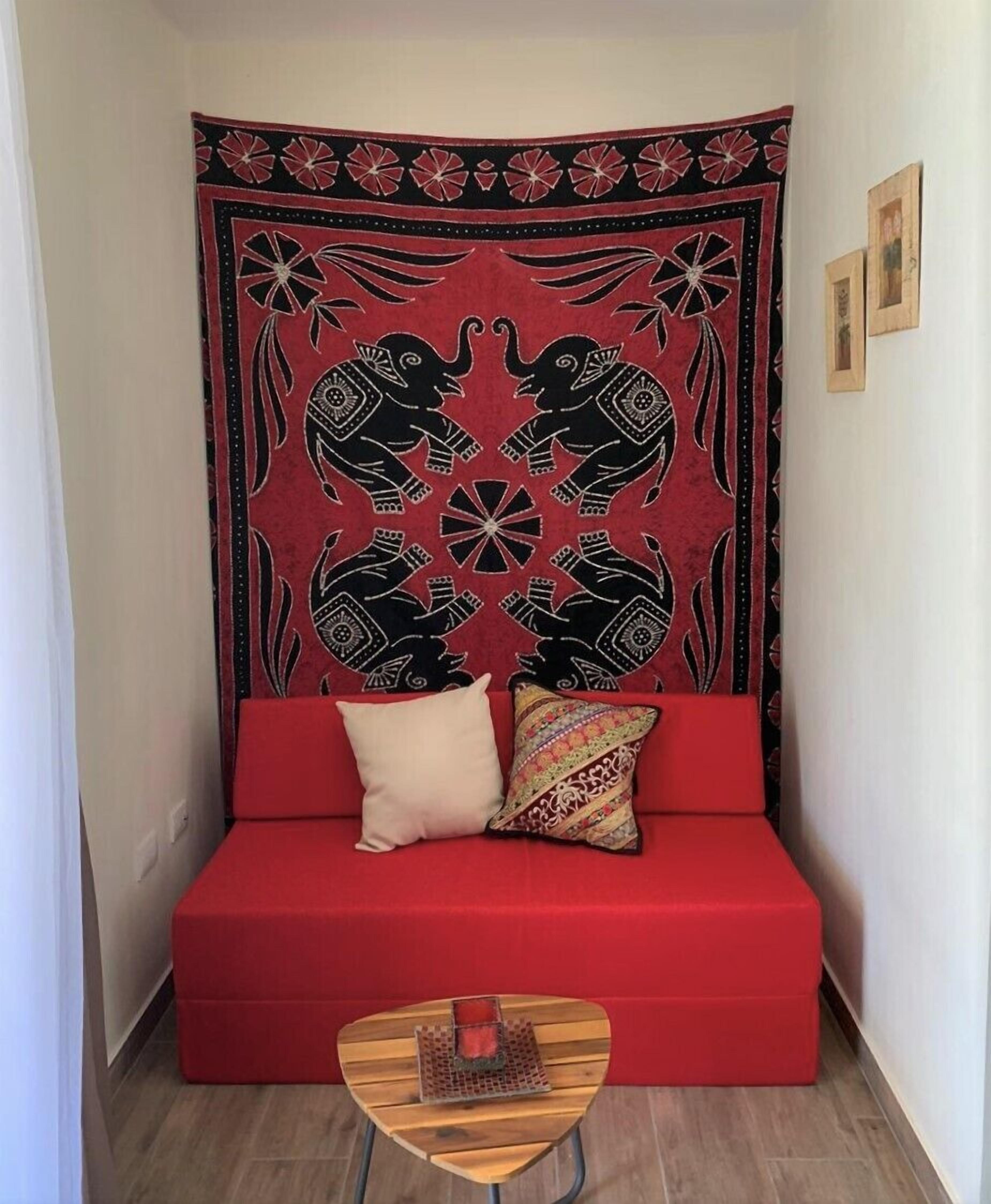 Um sofá vermelho com duas almofadas, uma mesa de centro de madeira e um tapeçário com padrões em preto e vermelho.