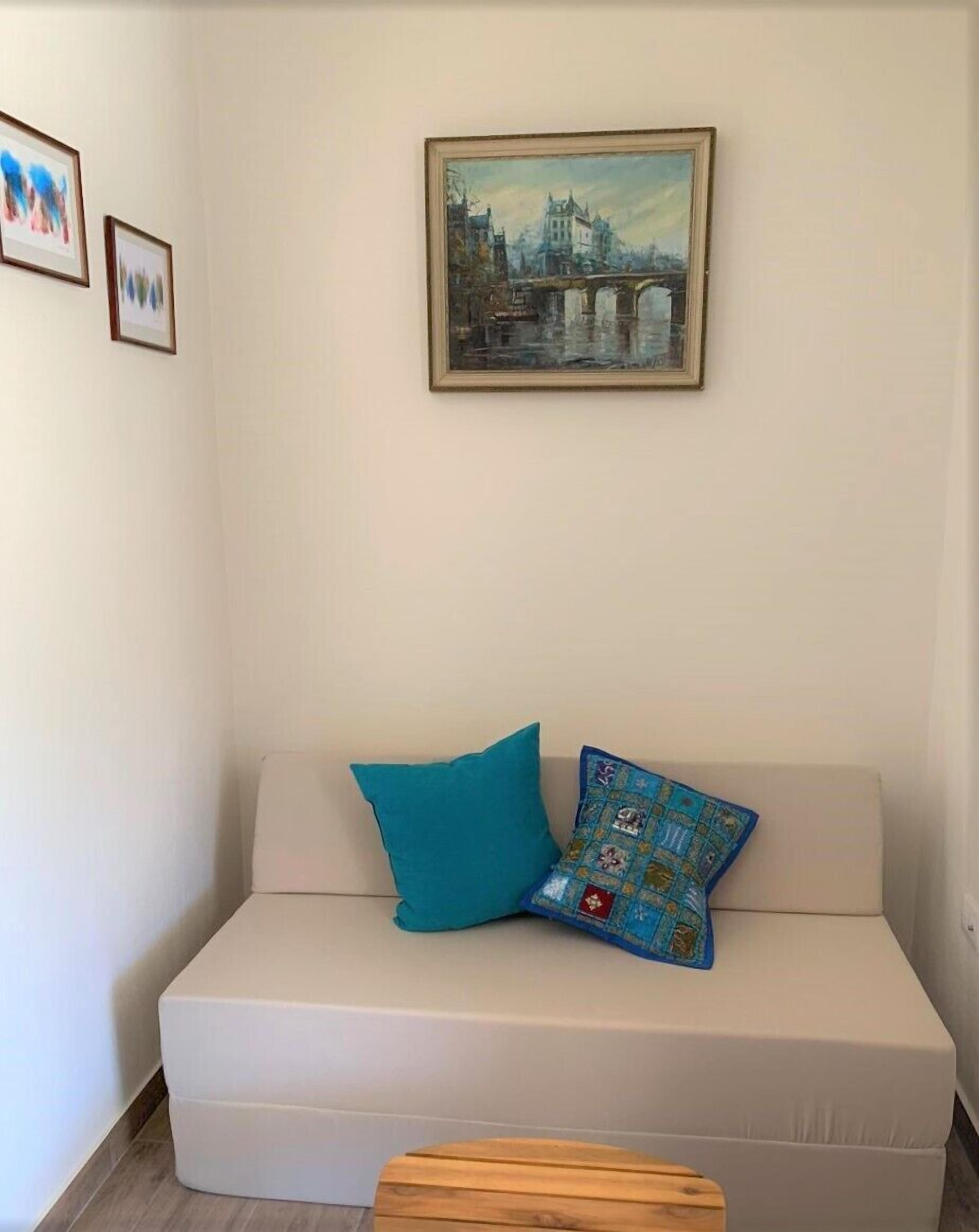 Een witte bank met twee kussens, daarboven een lijst met een schilderij en er voor een klein houten krukje.