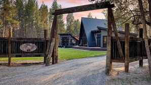 A rustic wooden gate with a sign, a gravel driveway, and a modern building with a sloped roof. - Newly Built Sauna Hot Tub Cold Punge Lazy River Glamp-house by Avantstay (La Pine)