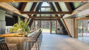 A spacious kitchen with a long counter, bar stools, and a large window overlooking a forest. - Newly Built Sauna Hot Tub Cold Punge Lazy River Glamp-house by Avantstay (La Pine)