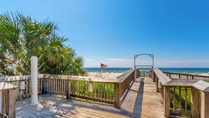 A wooden boardwalk leading to a beach with an American flag, surrounded by palm trees and clear skies.