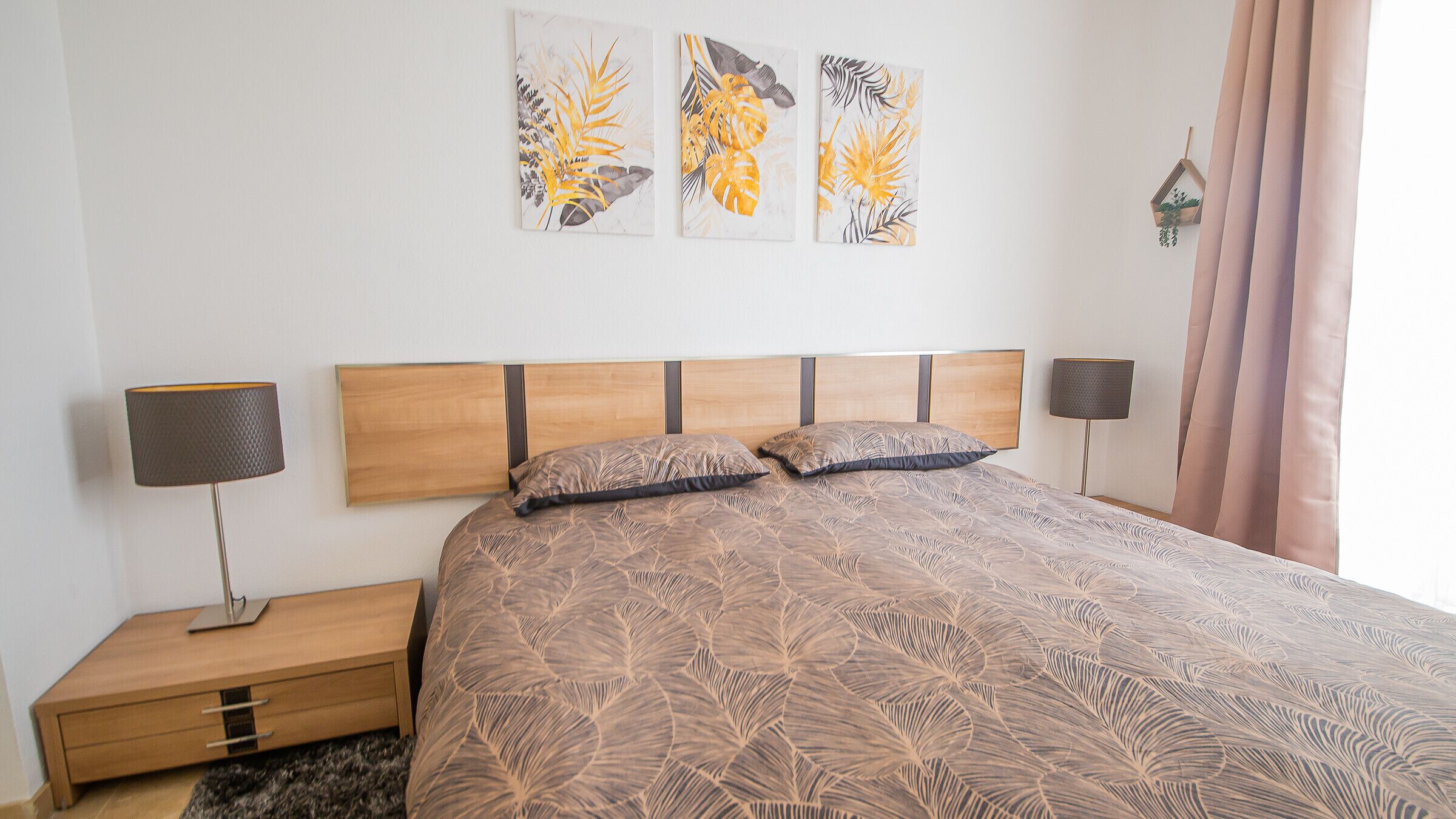 A bedroom with a wooden headboard, a patterned bedspread, two pillows, a nightstand, a lamp, and three wall art pieces.
