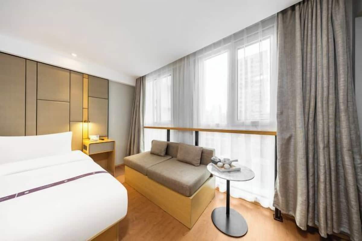 Habitació d'hotel moderna amb llit, sofà, taula rodona petita i finestra gran amb cortines.