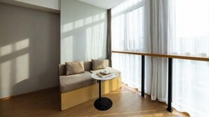 Una habitación moderna con suelo de madera, un sofá con cojines, una pequeña mesa redonda con un libro y una taza, y ventanas amplias con cortinas ligeras.