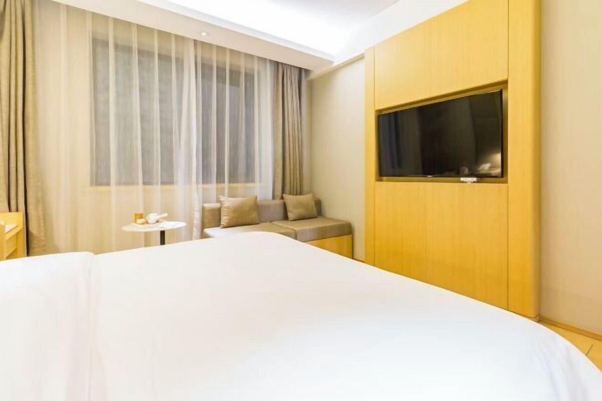 A hotel room with a large bed, a small sofa, a TV mounted on the wall, and a window with sheer curtains.