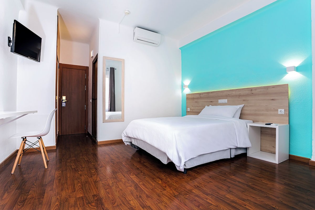 A modern hotel room with a wooden floor, a bed with white bedding, a desk with a chair, and a TV mounted on the wall.