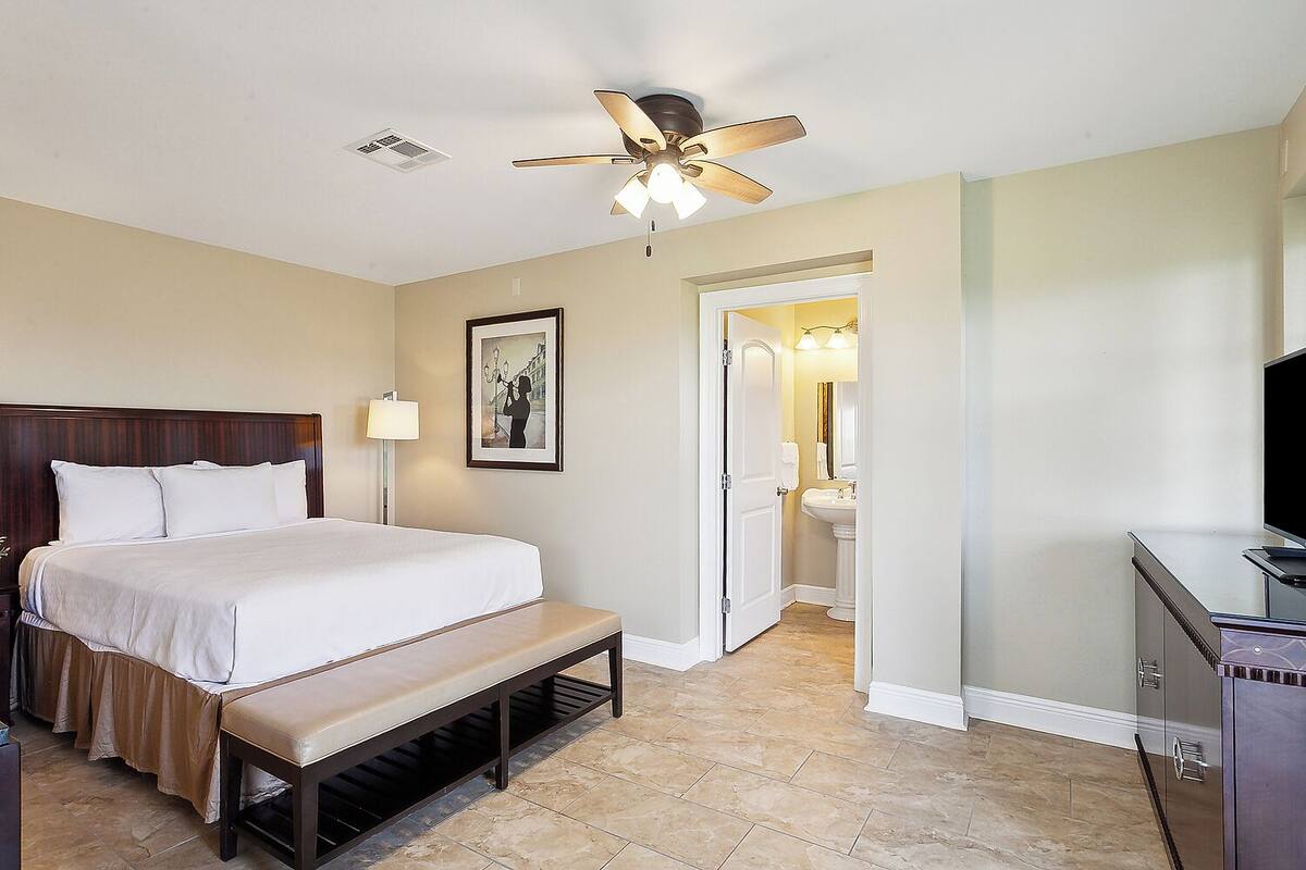 Habitación de hotel con cama, banqueta, ventilador de techo, cuadro enmarco, baño con lavamanos y espejo, y televisor sobre un mueble.