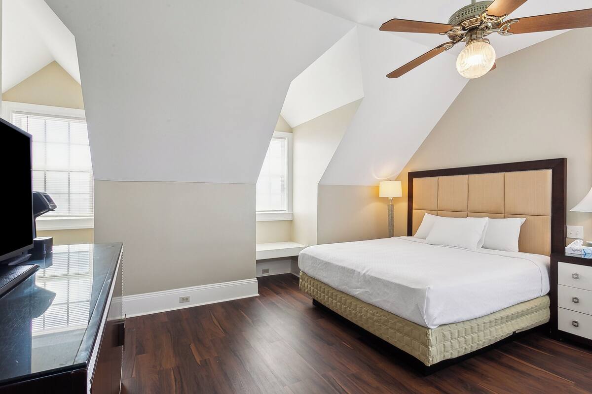 Un dormitorio con cama, escritorio, televisor, ventilador de techo y ventana.