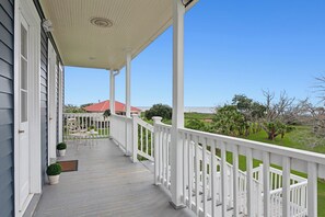 A covered porch with a white railing, potted plants, and a view of a red-roofed house and green landscape. - Elsa's Place Bed and Breakfast (Lafitte)