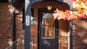 A brick building with a number "576" on the wall, a black door with a glass panel, and a small porch with a railing.