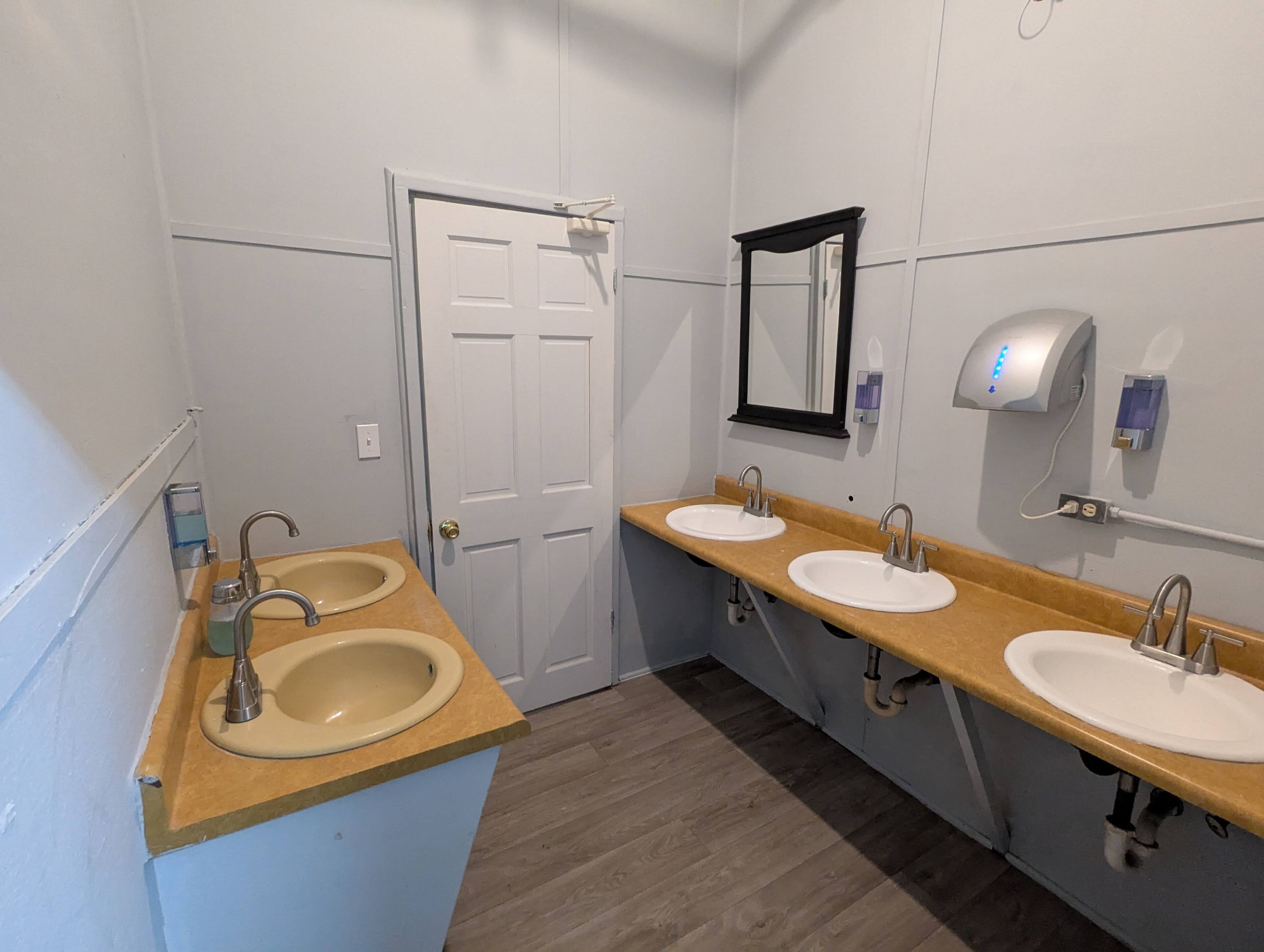 Een openbaar toilet met twee wasbakken, een spiegel, een zeepdispenser en een deur.