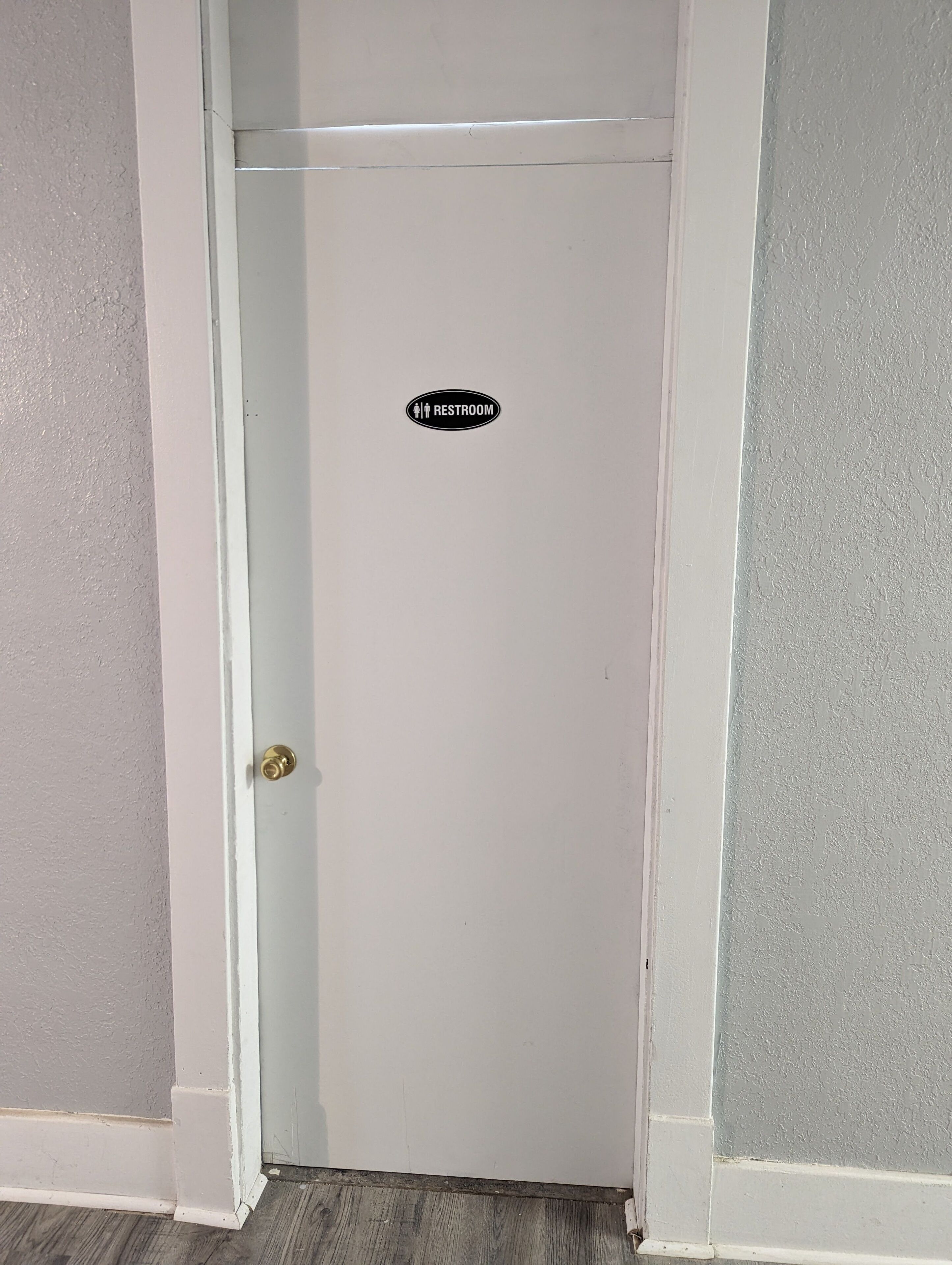 A white door with a restroom sign, a brass doorknob, and a wooden floor.