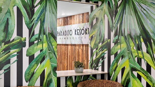 A room with a wooden slat wall, a sign reading "PARADISO RESORT KINGSCLIFF", and large green plant illustrations. - Paradiso 3 Bedroom Ground Floor 108 (Kingscliff)