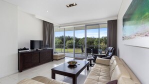 Ein modernes Wohnzimmer mit Sofa, Couchtisch, Fernseher auf einem Schrank und einem Balkon mit Blick auf Grünflächen.