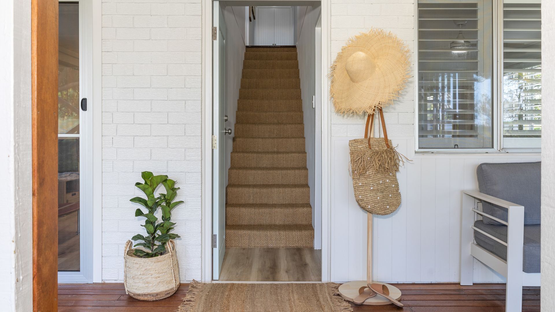 A staircase leading to a room with a white brick wall, a woven mat at the entrance, a woven basket with a plant, and a straw hat hanging on the wall.