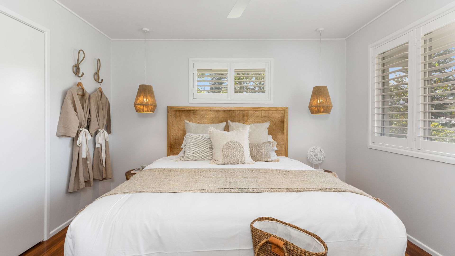 Un dormitorio con una cama grande, cabecera de madera, dos lámparas de pared, una ventana con persianas y una bata colgada.