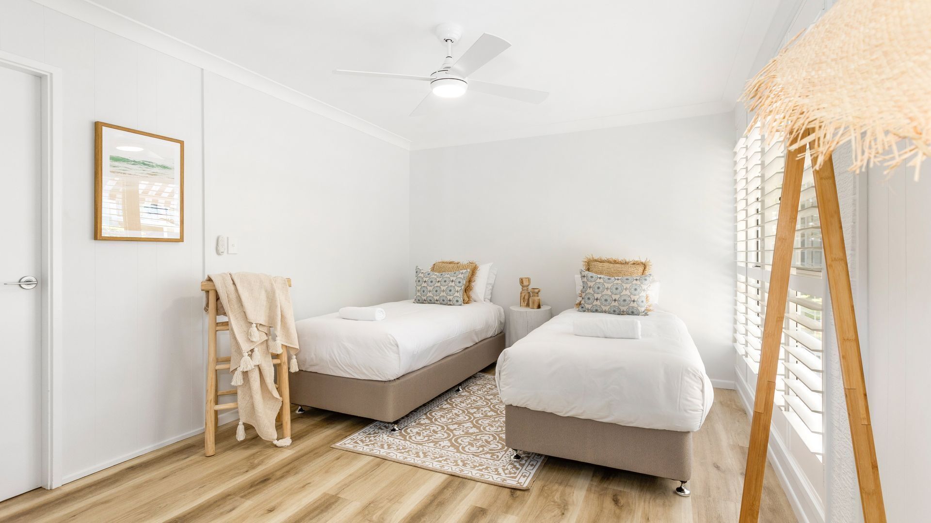 A bedroom with two beds, a ceiling fan, a wooden ladder, a patterned rug, and a window with blinds.