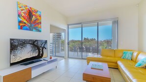 A modern living room with a flat-screen TV, a yellow leather sofa, and a large window offering a view of the outdoors. - Drift South 34 - Luxurious rooftop holiday apartment with lagoon pool views (Casuarina)