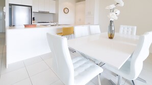A modern kitchen with white cabinetry, a stainless steel refrigerator, and a gas stove. There is a dining area with a white table and chairs, and a vase with white flowers on the table. - Drift South 34 - Luxurious rooftop holiday apartment with lagoon pool views (Casuarina)